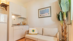 Cocina compacta con armarios blancos, un refrigerador blanco y un sofá color beige. En la pared, se observa un cuadro enmarcado, y una planta en maceta es visible.