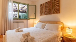 Una cama bien hecha con sábanas blancas y toallas enrolladas, una mesita de mimbre con una lámpara, y un ventanal con vistas a zonas verdes.