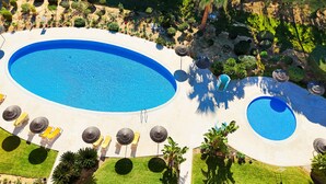 Vue aérienne d’un complexe hôtelier doté d’une grande piscine centrale, de plusieurs petites piscines secondaires et d’un jardin bien entretenu, agrémenté de palmiers et d’arbustes.