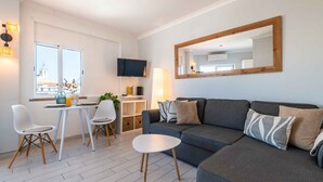 A modern living room with a grey sofa, white chairs, a small table, and a TV mounted on the wall. - Downtown Rooftop 1 Bedroom Apartment (Albufeira)