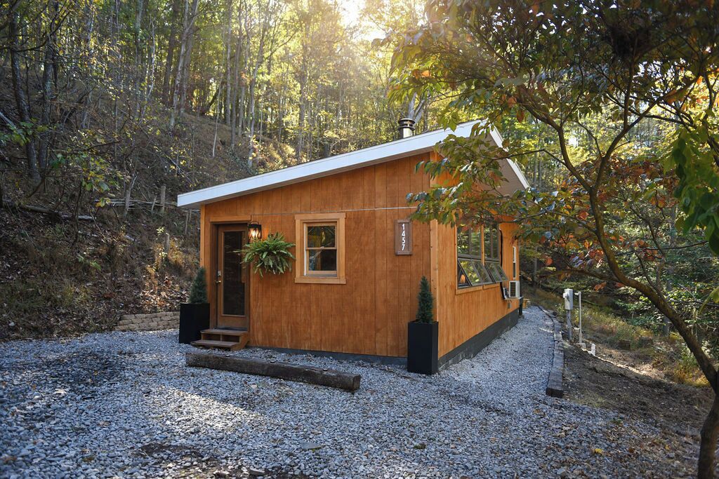 A small wooden cabin with a door, windows, and a chimney, surrounded by trees and gravel.