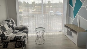 A room with a patterned sofa, a small round table, a white cabinet, and a view of a parking lot through a large window with blinds.