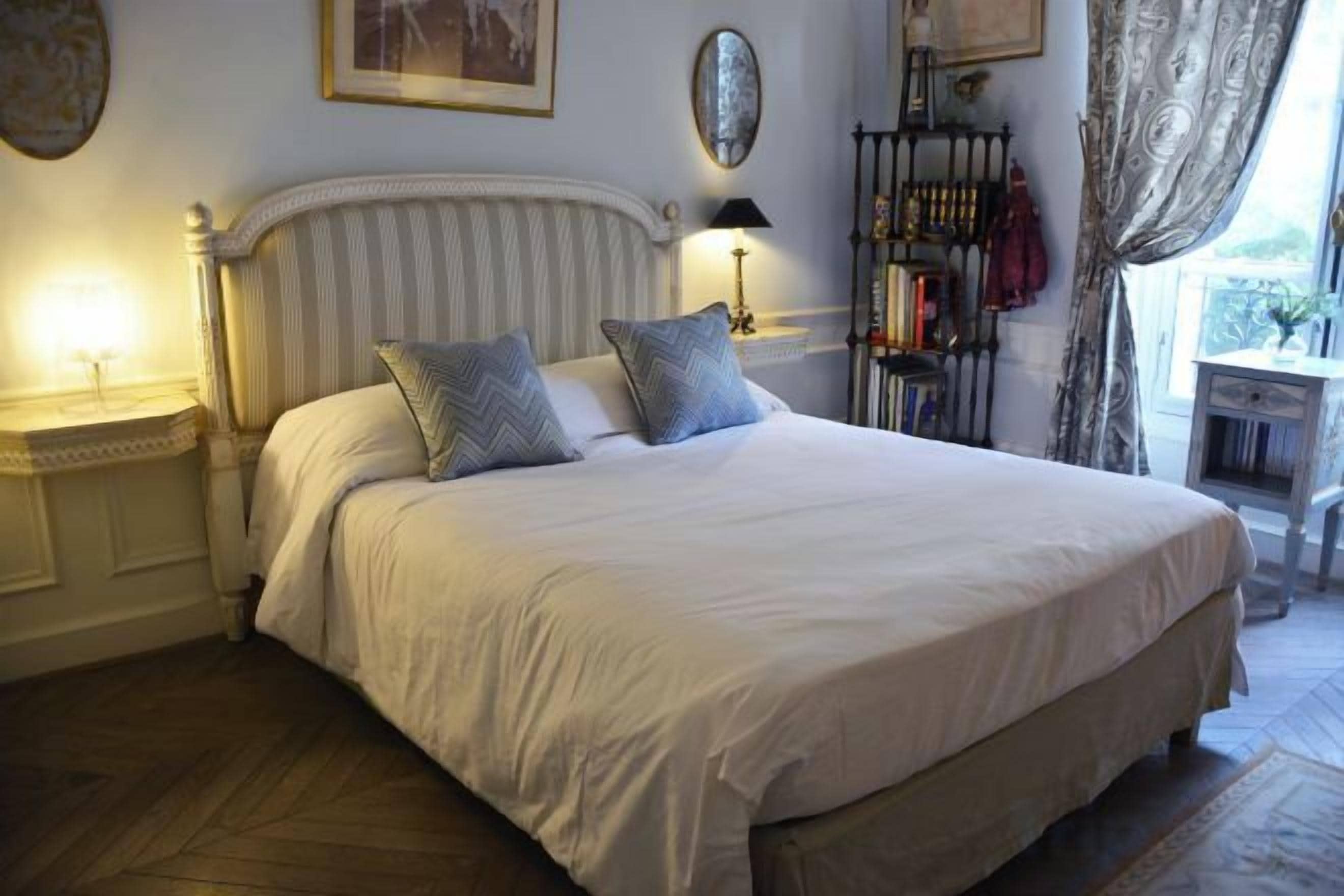 Een net opgemaakt bed met kussens, een nachtkastje met een lamp, en een raam met gordijnen.
