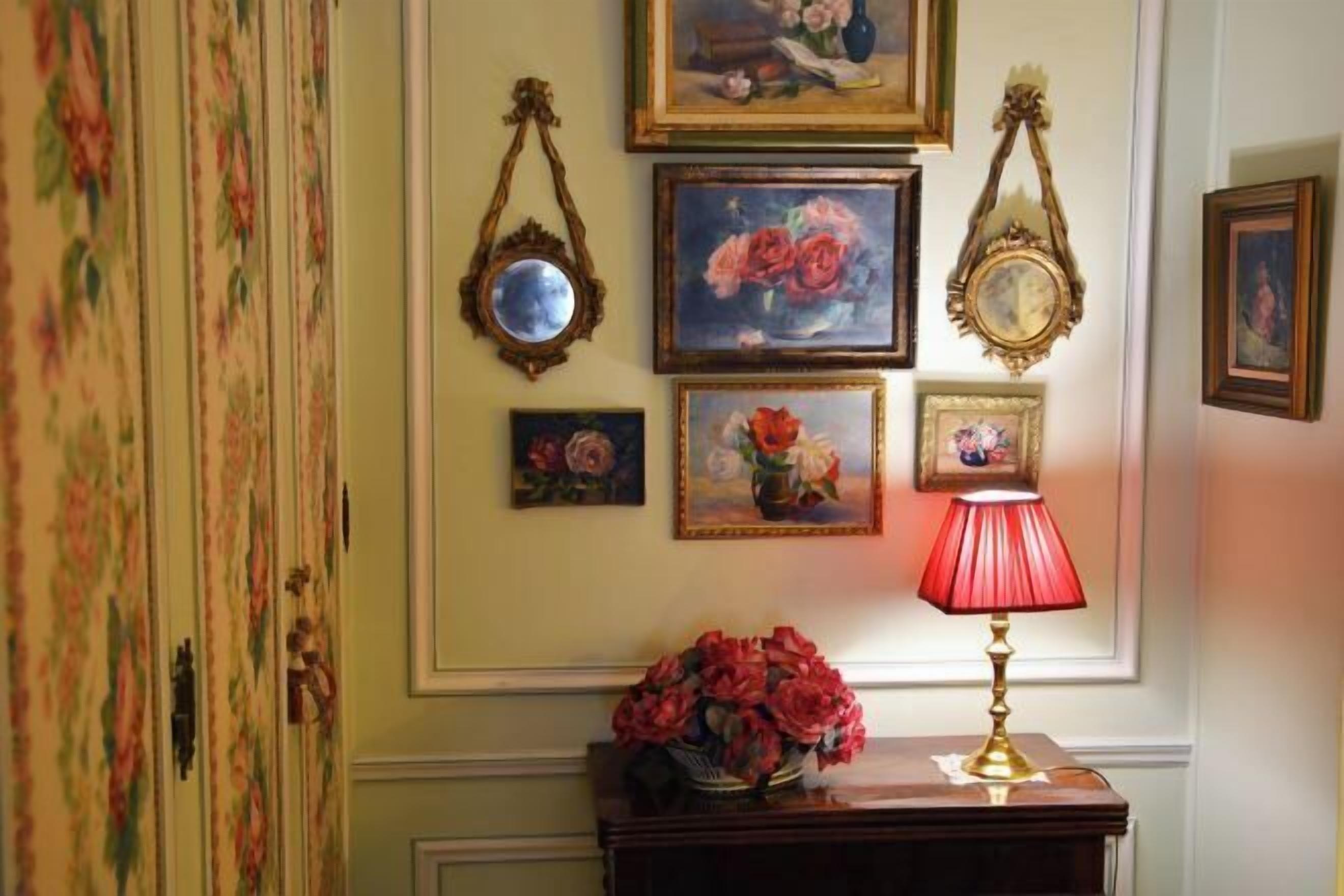 A wall with framed floral paintings, a mirror, and a table with a red lamp and flowers.
