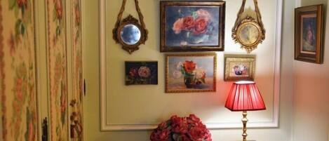 A wall with framed floral paintings, a mirror, and a table with a red lamp and flowers.