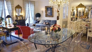 A room with a glass-top dining table, clear chairs, a red armchair, a sofa, and a chandelier. - Elegant 3 Bedroom Apt Near Eiffel Tower - Alma (Paris)