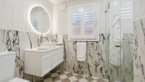 A bathroom with a white vanity, a round mirror, a window with blinds, and a shower area. - Spencer House in Leura (Leura)