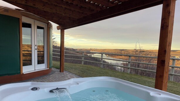 Un jacuzzi con agua corriendo, una terraza de madera, una puerta de cristal y vistas a un río y colinas.