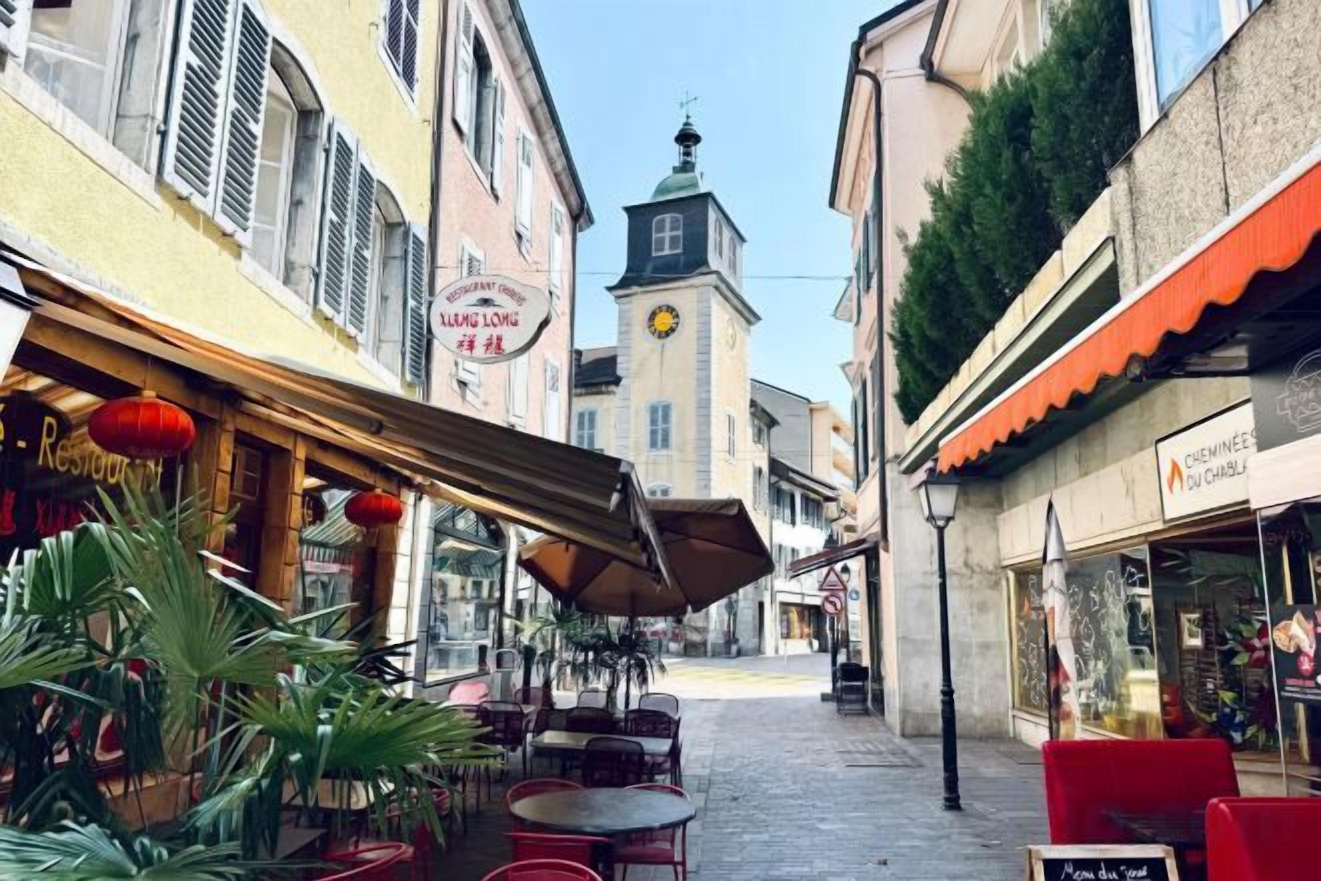 A narrow street with outdoor seating, a clock tower, and various shops.