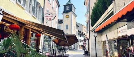A narrow street with outdoor seating, a clock tower, and various shops.