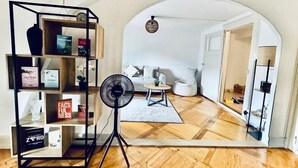 Une pièce avec un plancher en bois, un ventilateur noir, un canapé blanc, une petite table et une bibliothèque.