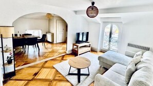 Un vaste espace de vie doté d’un parquet en bois, d’une table basse ronde, d’un canapé gris, d’un téléviseur et d’un coin repas avec une table et des chaises.