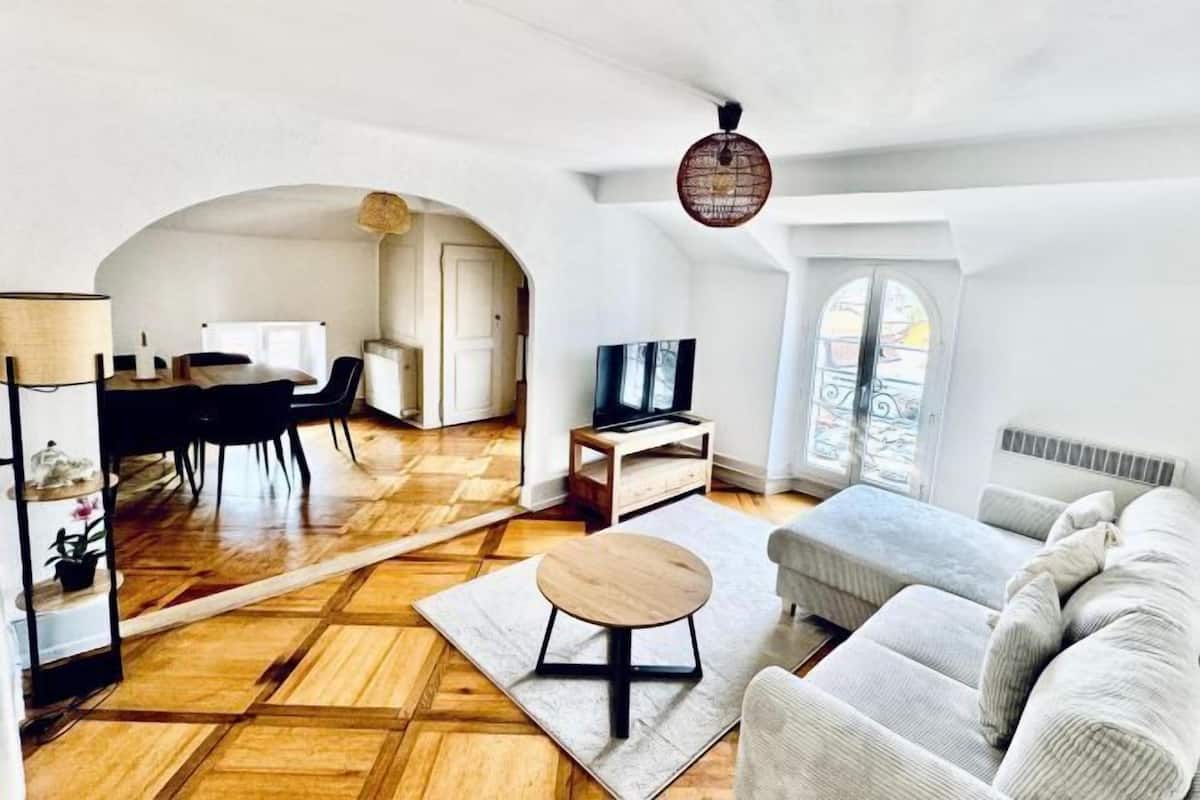 Amplia sala de estar con piso de parquet de madera, una mesa redonda para café, un sofá gris, un televisor y un comedor con mesa y sillas.
