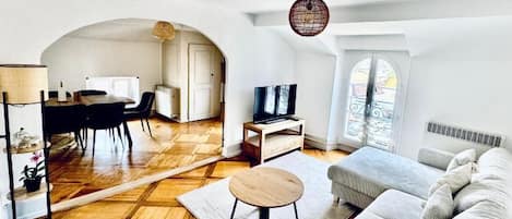 A spacious living area with a wooden parquet floor, a round coffee table, a grey sofa, a television, and a dining area with a table and chairs.
