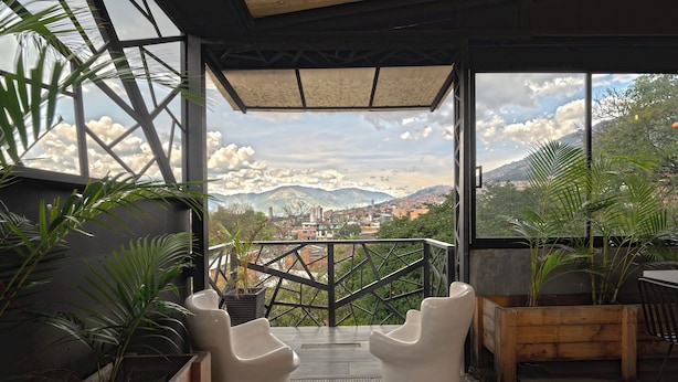 A balcony with two white chairs, potted plants, and a view of a cityscape and mountains. - Luxury Suite - Rooftop, Jacuzzi & Balcony (Medellín)