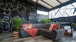 A modern living room with a sectional sofa, a large abstract wall mural, and a view of the outdoors through a geometric window structure. - Luxury Suite - Rooftop, Jacuzzi & Balcony (Medellín)