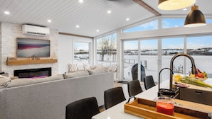 Ett modernt vardagsrum med en öppen spis, ett stort fönster med utsikt över ett snötäckt landskap och en matplats med ett bord och stolar.