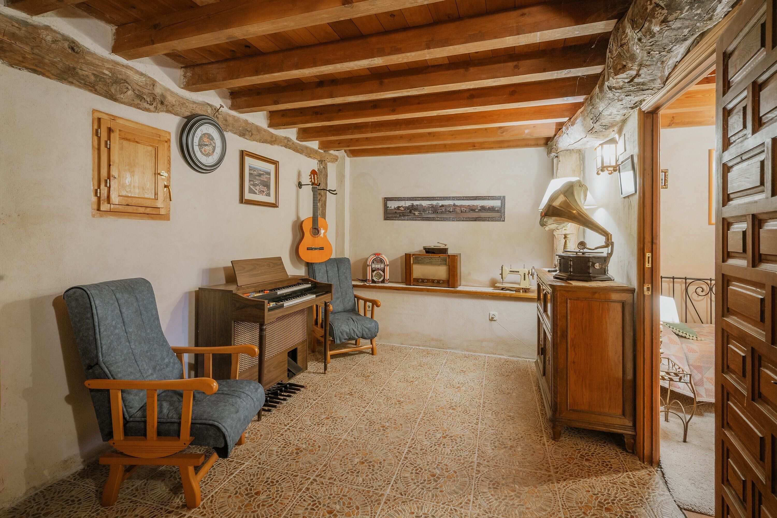 A room with a wooden ceiling, a piano, a guitar, a radio, and a bed with a patterned bedspread.