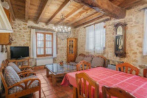 A rustic room with a wooden ceiling, stone walls, a dining table with chairs, a sofa, a television, and a clock.