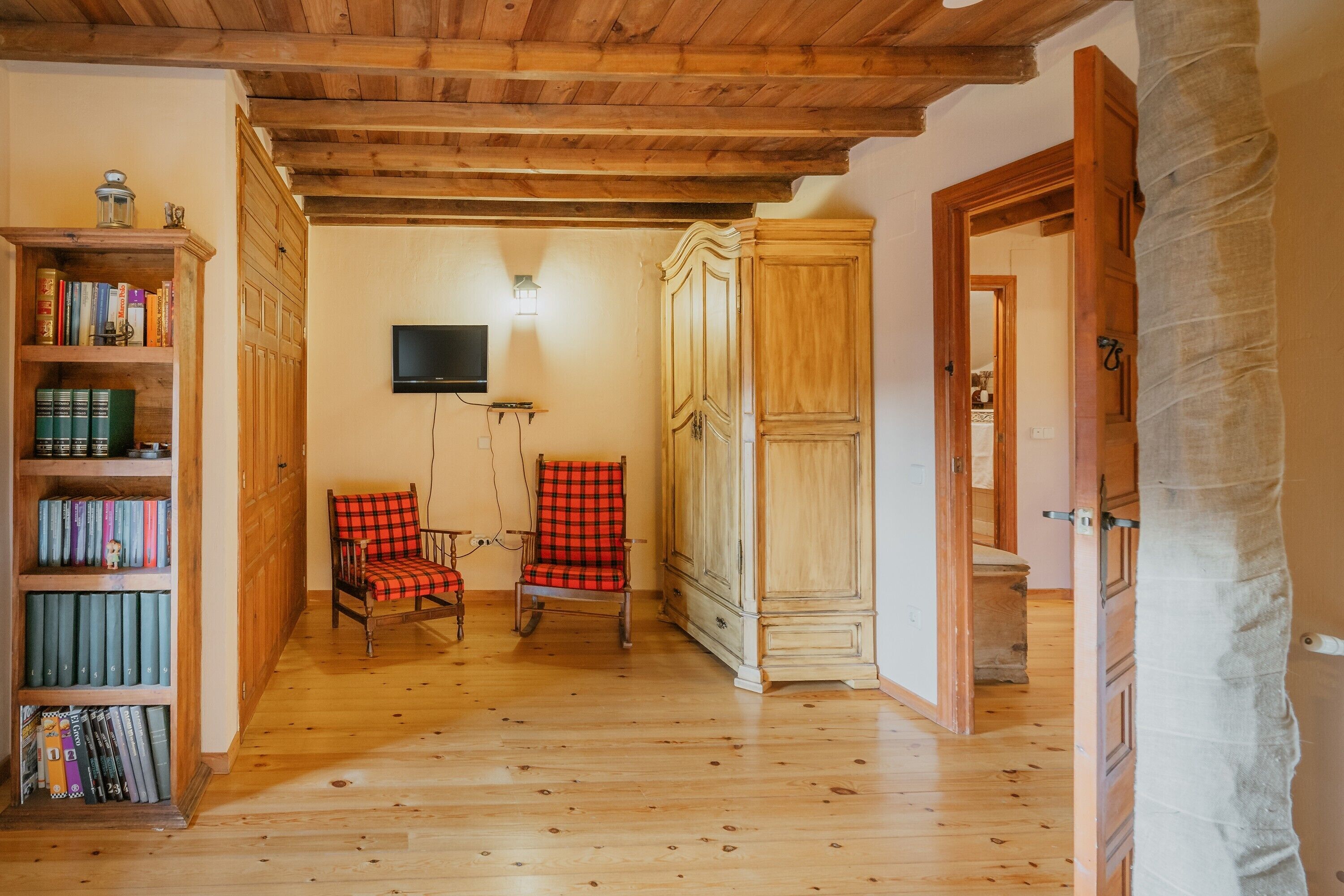 Una habitación con suelo de madera, una estantería, un sillón rojo a cuadros y un televisor montado en la pared.