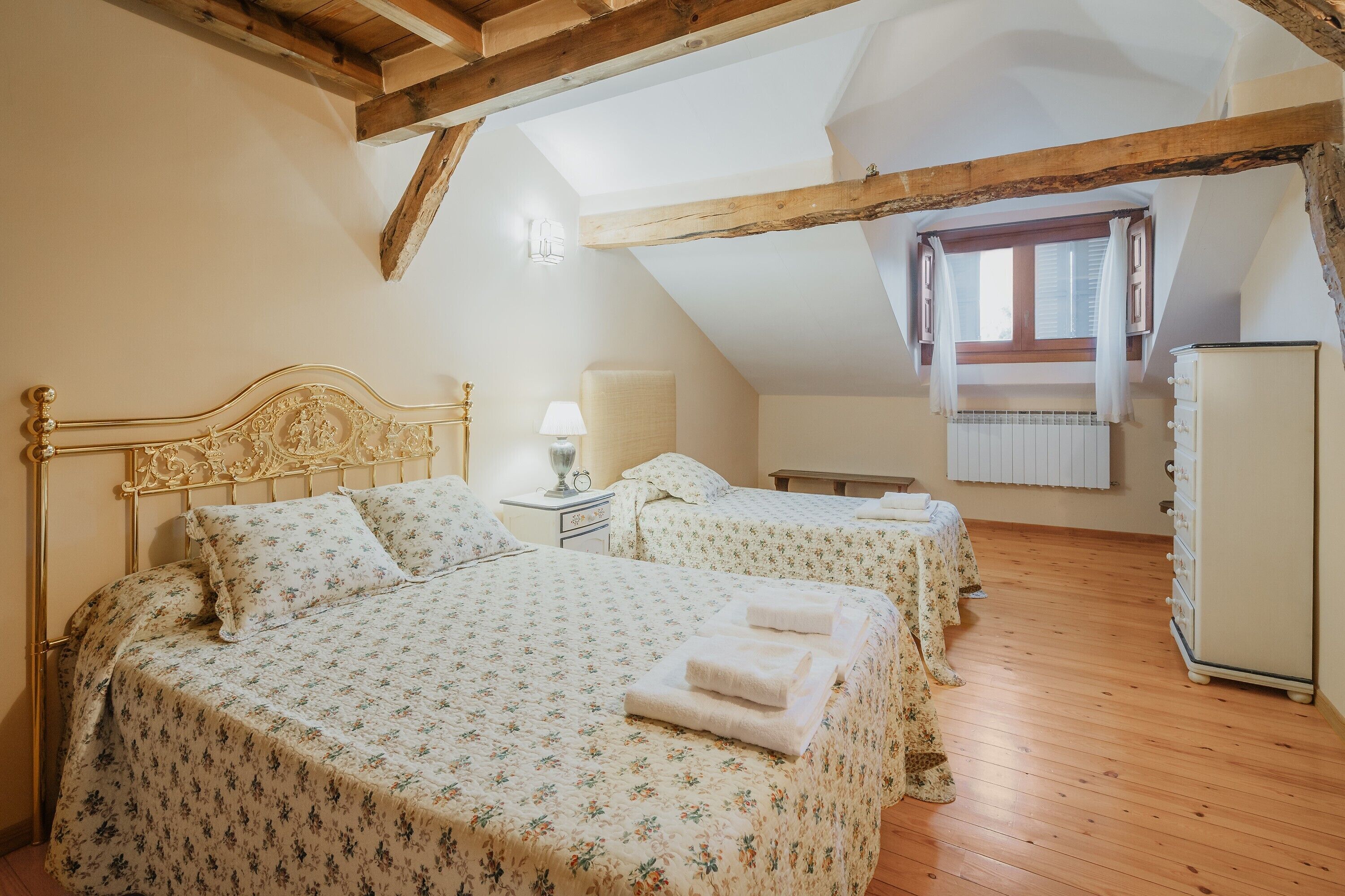 A bedroom with two beds, wooden beams, a window with curtains, a bedside table with a lamp, and a wardrobe.