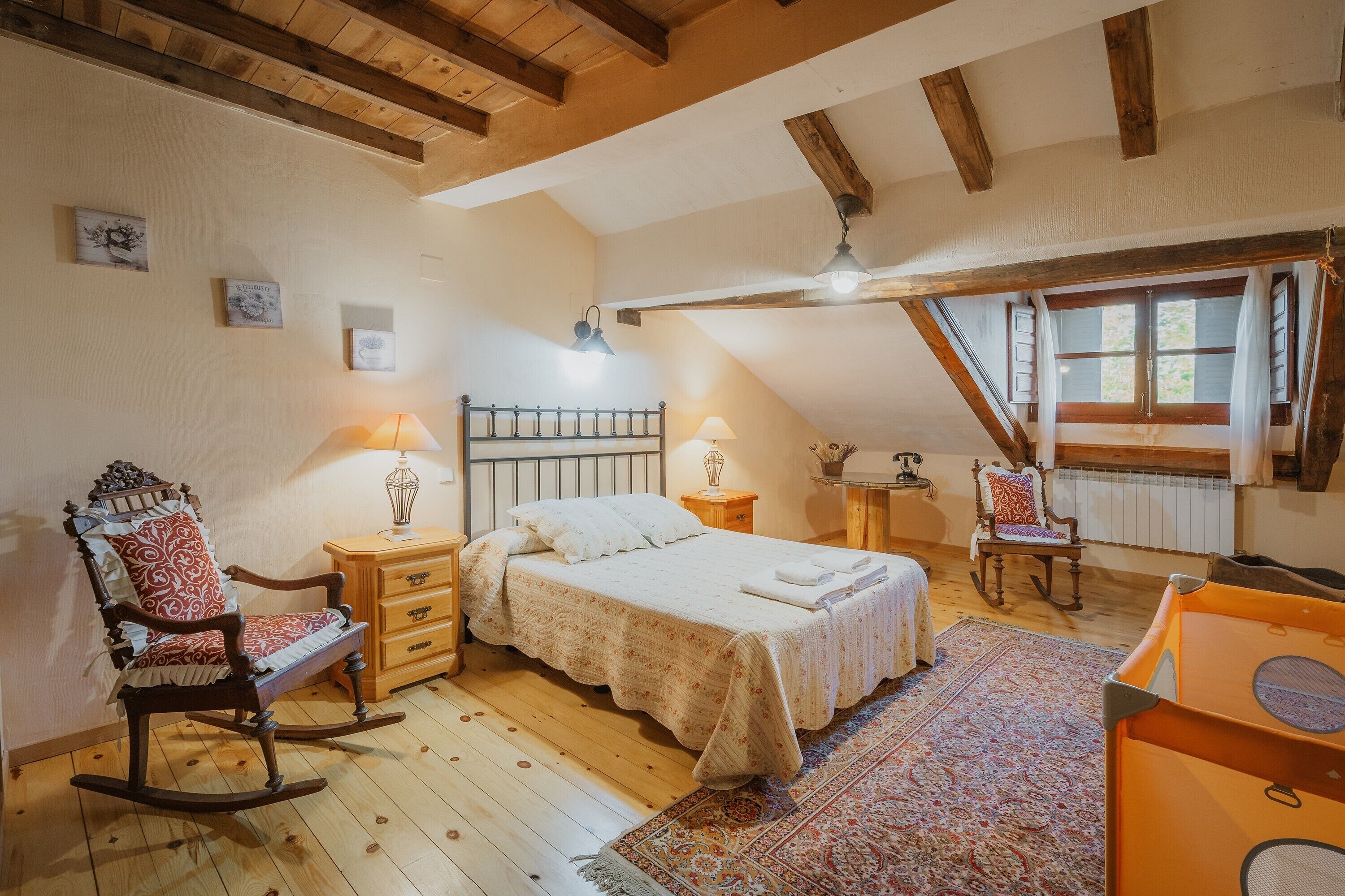 Un dormitorio con cama, sillón mecedor, mesitas de noche y una ventana con cortinas.