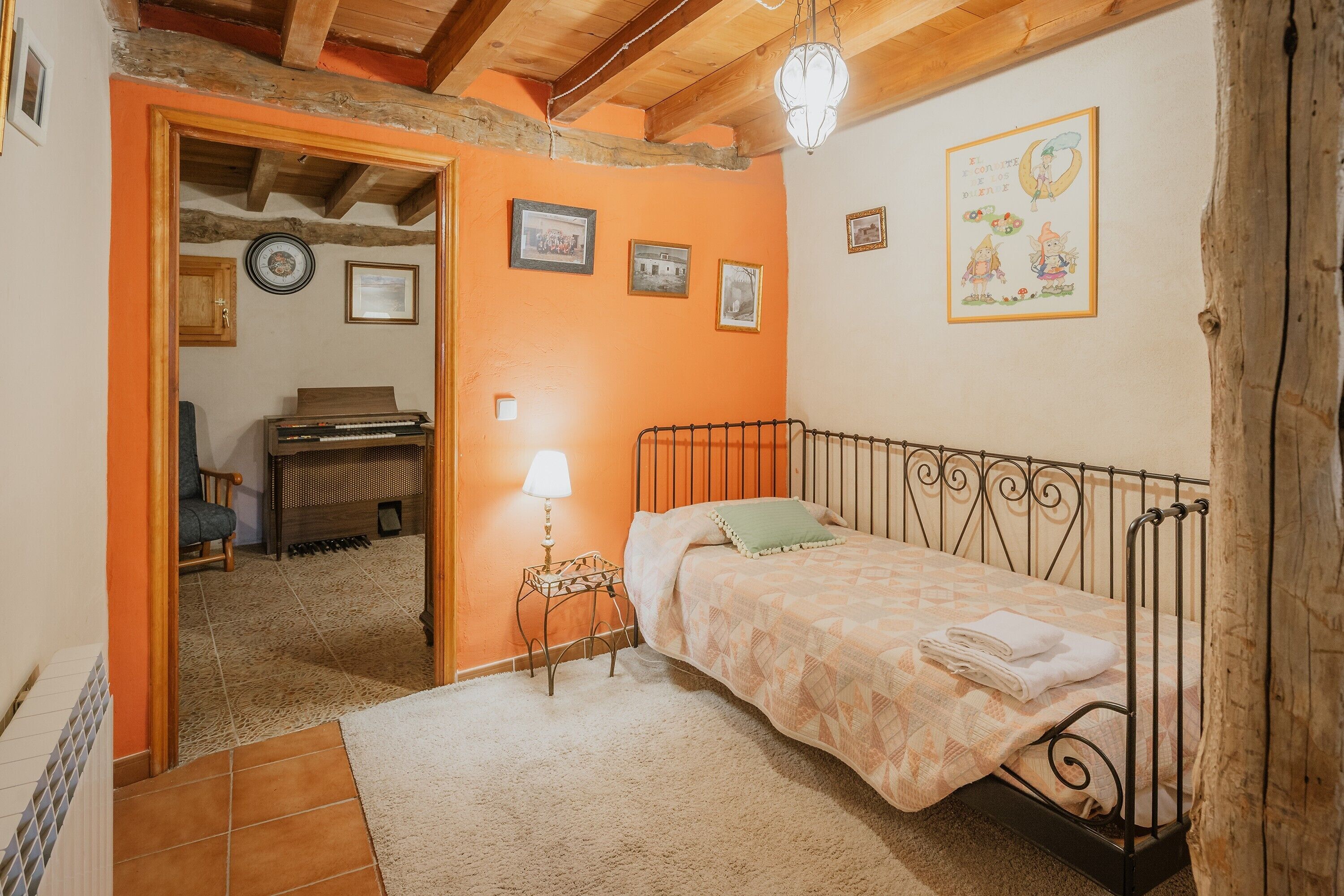 A bedroom with a bed, a nightstand, a lamp, a piano, and framed pictures on the wall.