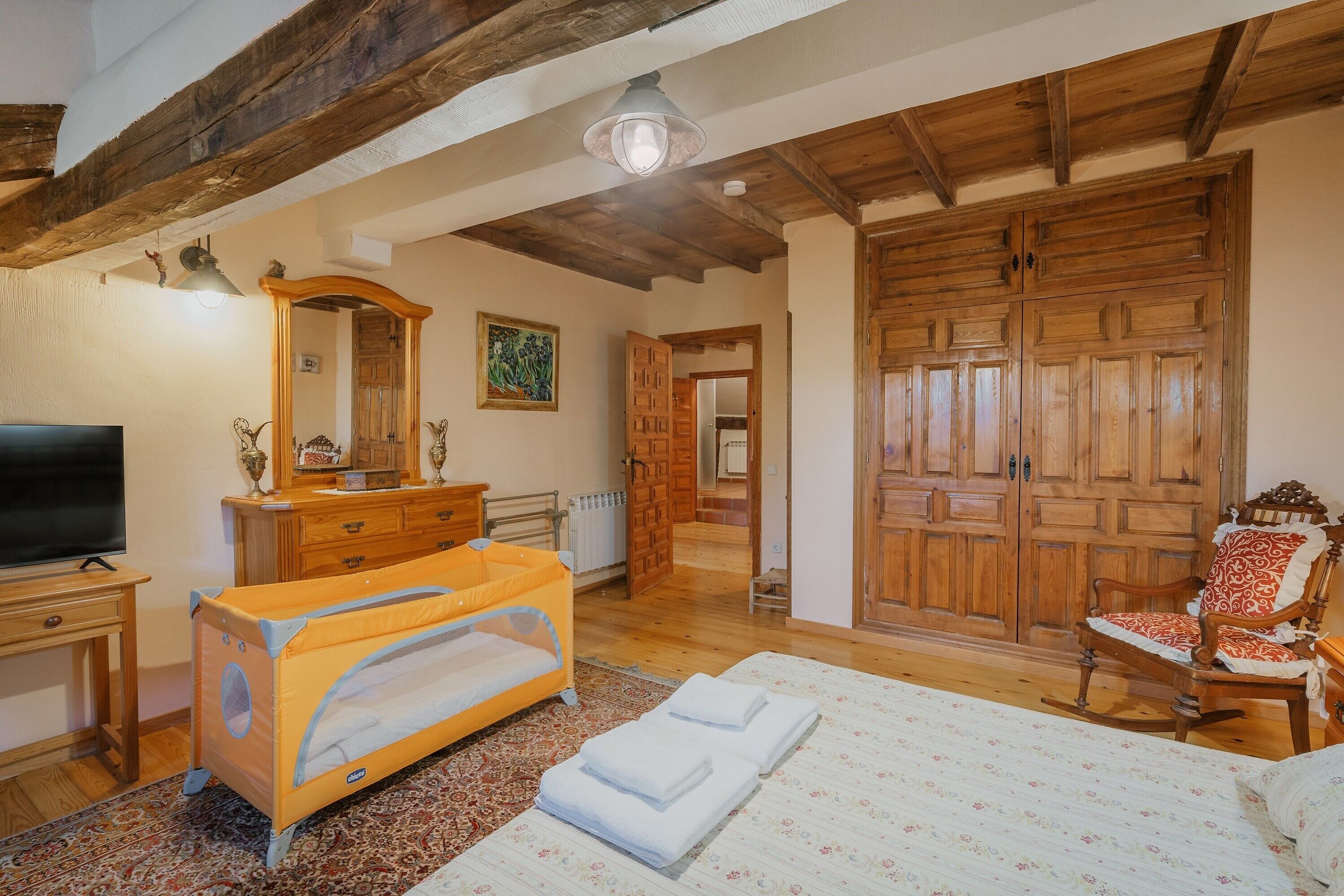 A room with a bed, a wooden wardrobe, a television, a crib, and a rocking chair.
