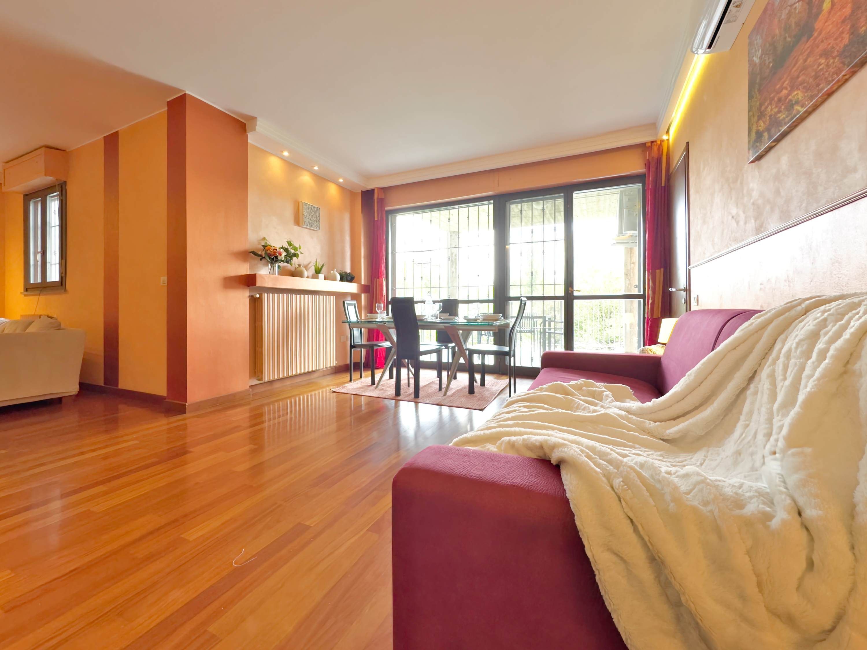 A spacious living area with a wooden floor, a red sofa, a dining table with chairs, and a large window providing natural light.