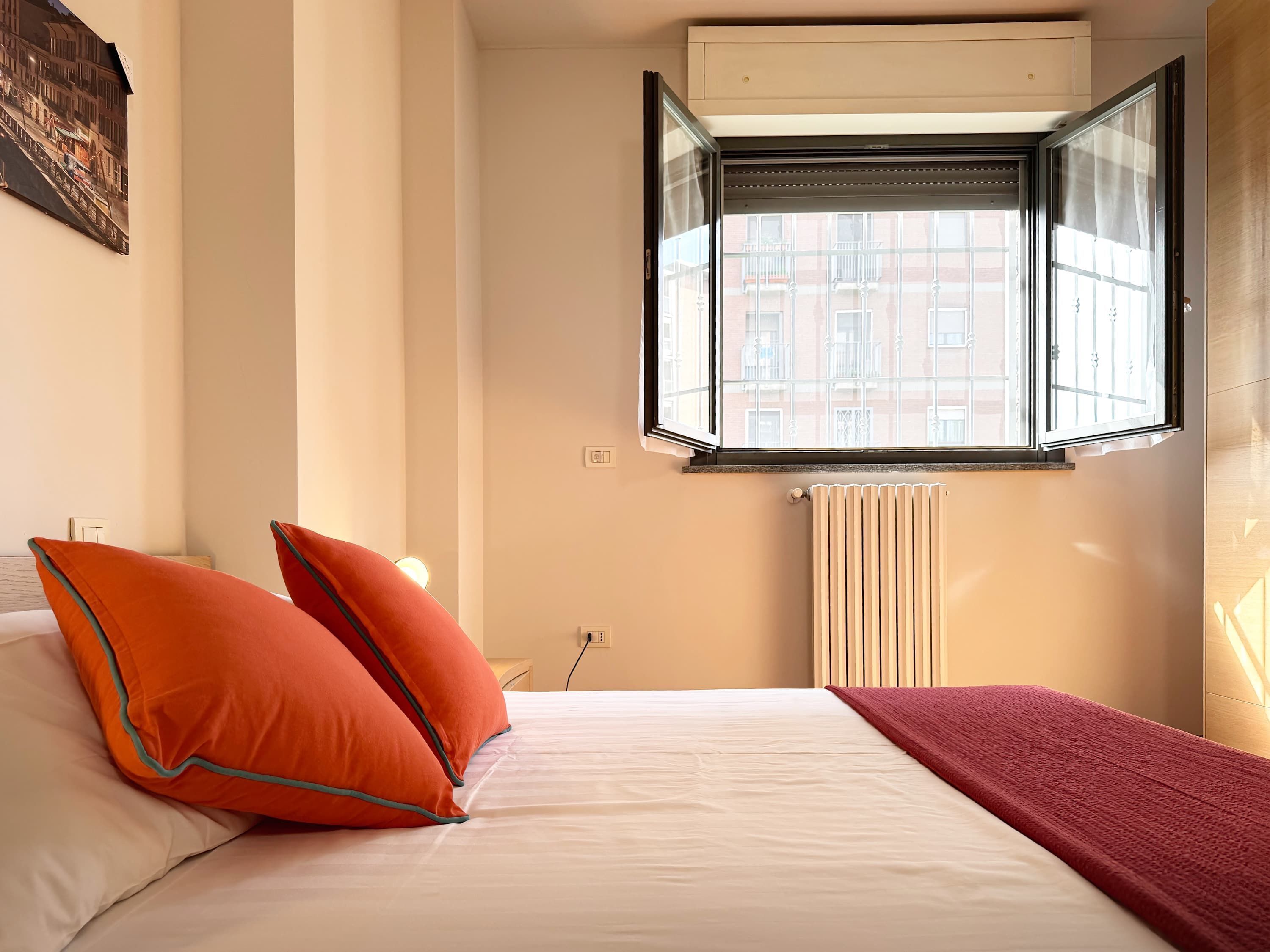 Un dormitorio con una cama, dos almohadas naranjas, una manta roja, una ventana con vistas a edificios y un radiador.
