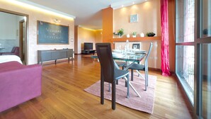 Una habitación de hotel moderna con cama, escritorio, televisión y zona de comedor con mesa y sillas de vidrio.