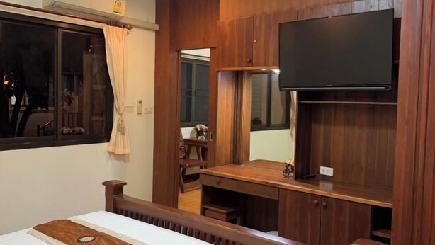 A room with a bed, a TV mounted on a wooden wall, a window with curtains, and a wooden desk. - Rommaiplaitawan hotel & resort (Nakhon Ratchasima)