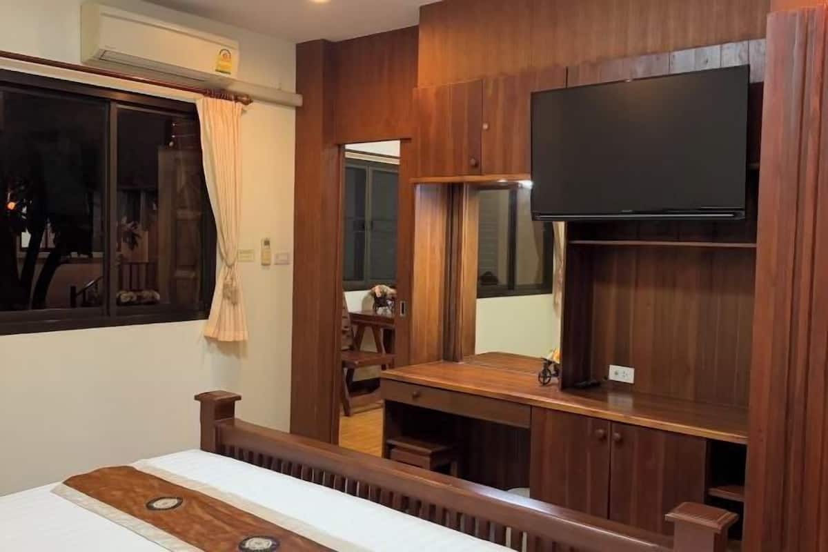 Ein Zimmer mit einem Bett, einem an einer Holzwand montierten Fernseher, einem Fenster mit Vorhängen und einem Holztisch.
