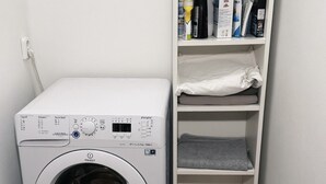 A compact, white front-loading washing machine next to a white shelving unit with various items on it. - Bright and Cozy 40 m² Apartment at Prague 7 (Prague)
