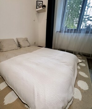 Une chambre à coucher avec un grand lit, une table de chevet, une étagère fixée au mur et une fenêtre ornée de rideaux légers.