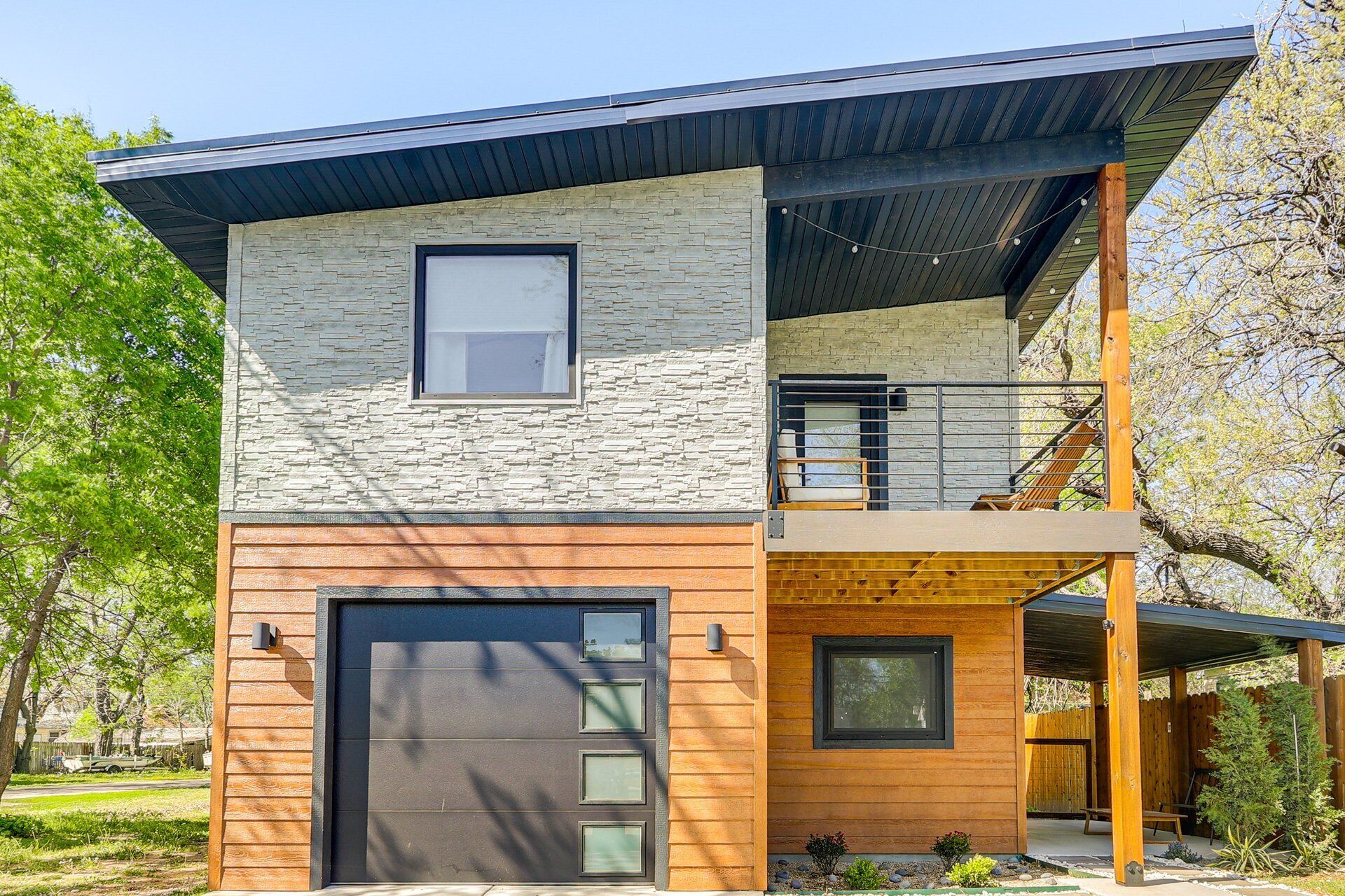 Une maison moderne à deux étages, dotée d’une façade en pierre, d’une terrasse en bois et d’un espace extérieur couvert.