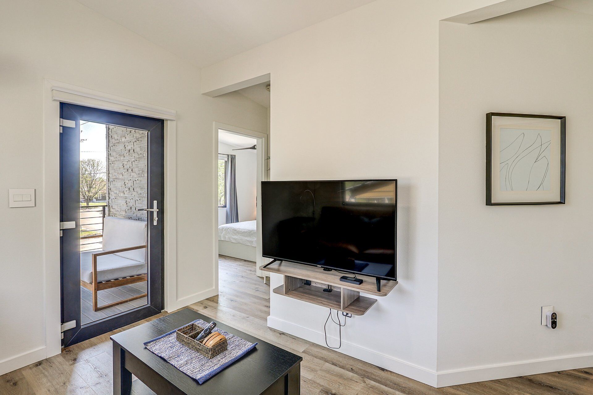 Un salon moderne avec un téléviseur à écran plat fixé à un mur blanc, une petite table sur laquelle se trouvent une télécommande et un objet décoratif, et une porte donnant sur un balcon.