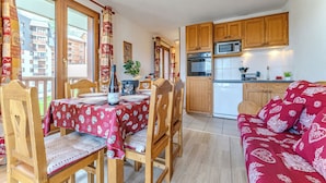 A cozy living room with a red patterned sofa and wooden chairs, a dining area with a table set for two, and a kitchen with wooden cabinets and modern appliances. - Chalet Niverolles (Les Belleville)