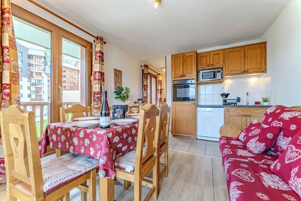 Un salon chaleureux avec un canapé rouge à motifs et des chaises en bois, un coin repas avec une table dressée pour deux personnes, et une cuisine dotée de meubles en bois et d’appareils électroménagers modernes.