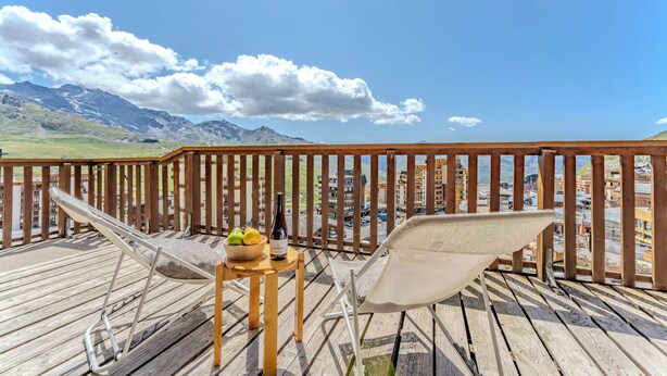 A wooden deck with two white mesh lounge chairs, a small wooden table with a bowl of fruit and a bottle of wine, and a view of mountains and buildings in the background. - Chalet Niverolles (Les Belleville)