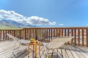Une terrasse en bois avec deux chaises longues blanches en tissu ajouré, une petite table en bois sur laquelle se trouve un saladier rempli de fruits et une bouteille de vin, et une vue sur des montagnes et des bâtiments en arrière-plan.
