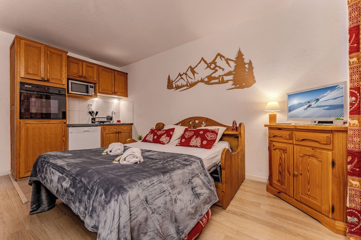 A bedroom with a bed, wooden cabinets, a TV, and a kitchen area.