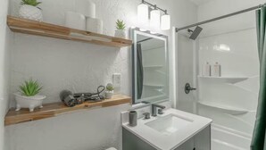 Ein Badezimmer mit weißem Waschbecken, einem Spiegel und einer Dusche. Holzregale mit Pflanzen und Pflegeprodukten sind ebenfalls vorhanden.