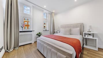 A modern bedroom with a large bed, a bedside table, a radiator, and a window with a view of buildings.