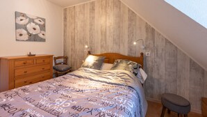 Een slaapkamer met een schuine kap, een houten hoofdbord, een bed met beddengoed met een patroon, een nachtkastje, een dressoir en een wandlamp.