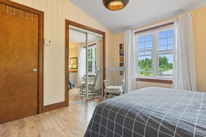 A bedroom with a bed, a chair, a window with curtains, a door, and a mirror. - Kamouraska, Lac et nature (Mont-Carmel)
