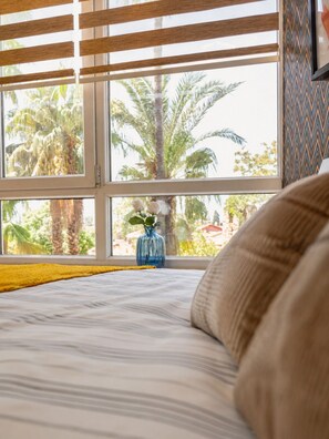 A bedroom with a bed, a window with blinds, a vase with flowers, and a view of palm trees outside. - Maxihomes Hadrian Amber (Antalya)