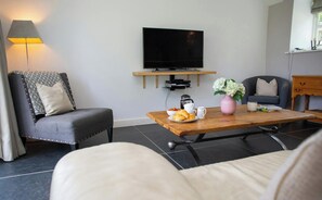 A modern living room with a flat-screen TV mounted on a wooden shelf, a comfortable sofa, a coffee table with a croissant and flowers, and a lamp.