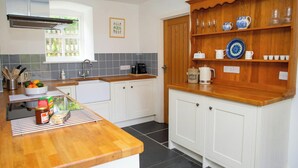A kitchen with white cabinets, a wooden countertop, a built-in shelf with glassware, and a window with a view of greenery. - The Dovecote, Llechryd (Llechryd)
