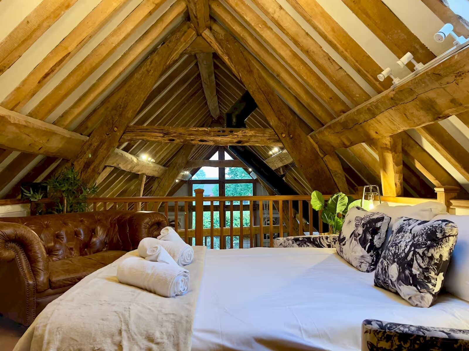 A cozy attic bedroom with a wooden ceiling, a bed with white bedding, a brown leather sofa, and a view of greenery outside.